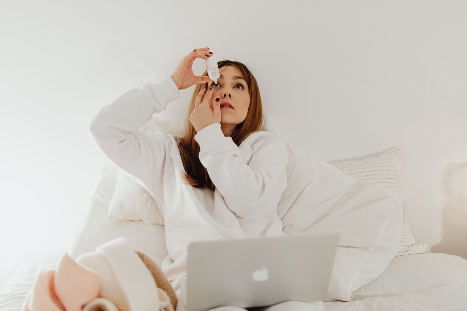 A woman applying eye drops while resting in bed with a laptop, symbolizing relaxation and self-care at home.